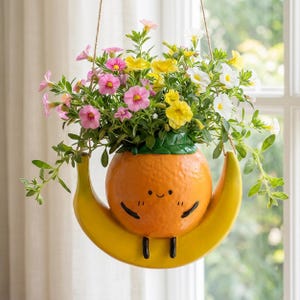 May include: A hanging planter shaped like a smiling orange fruit sitting on a yellow banana crescent. The orange has a textured surface, black painted eyes, a small smile, and black legs. It is filled with a variety of pink, yellow, and white flowers.