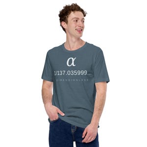 May include: A slate blue short-sleeved t-shirt featuring a white graphic print of the Greek letter alpha, the numerical value 1/137.035999, and the word DIMENSIONLESS printed in a smaller font underneath the number on the chest area.