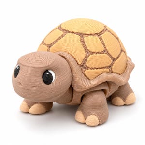 May include: A 3D-printed tortoise figurine featuring a light brown head and limbs with a tan shell. The surface displays visible horizontal layer lines from the printing process. The tortoise has large, round, black eyes with white pupils.