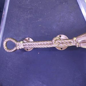 May include: A gold-toned metal door handle featuring a textured rope design. The handle includes a circular loop at one end, a flared bell-shaped terminal at the other, and two circular mounting plates with pre-drilled holes for installation.