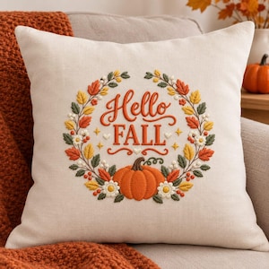 Op de afbeelding: Een vierkant gebroken wit sierkussen met een geborduurd kransontwerp. De tekst in het midden luidt Hello FALL in oranje letters. De krans bevat oranje en gele bladeren, witte bloemen en een oranje pompoen in het onderste gedeelte.