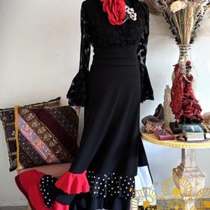 May include: A black long-sleeved dress featuring a textured bodice, bell sleeves, and a high waist. The skirt includes layered ruffles in red, black with white polka dots, and white. Two large red fabric roses are attached to the neckline.