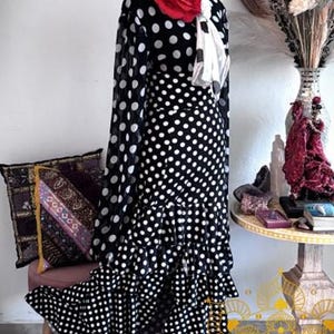 Puede incluir: Un vestido negro con lunares blancos que presenta mangas largas transparentes, cintura alta y una falda con volantes escalonados. La prenda está adornada con dos grandes rosas de tela roja en el escote y un pañuelo de lunares blancos al cuello.