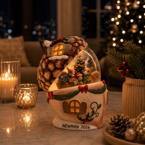 May include: A decorative acorn-shaped snow globe featuring a Christmas tree and three squirrels inside. The base is cream-colored with green garland, red bows, and the text NEWMAN 2026. The top resembles a textured brown acorn cap with a lit window.