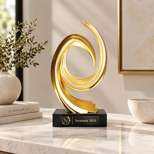 Può includere: Una scultura astratta color oro con linee curve e fluide montata su una base rettangolare nera. La base presenta una targhetta con la lettera N all'interno di un emblema circolare e il testo Newman 2026 stampato in un carattere bianco senza grazie.