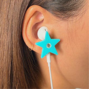 May include: A bright turquoise star-shaped earbud cover featuring a small, clear, circular rhinestone at its centre. The accessory is attached to a white wired earbud, which is positioned inside the ear canal, providing a decorative accent.