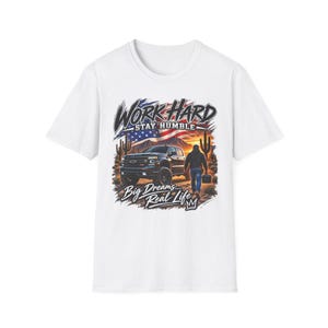 Puede incluir: Una camiseta blanca con un estampado gráfico de una camioneta negra, una persona cargando una caja de herramientas y cactus saguaro en un paisaje desértico bajo una bandera estadounidense. El texto dice WORK HARD STAY HUMBLE y Big Dreams Real Life.