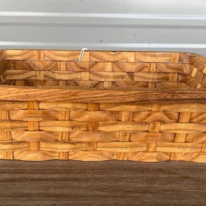 Op de afbeelding: Een rechthoekige, geweven houten mand met een lichtbruine afwerking. De mand heeft een stevige, vierkante vorm met een geweven patroon en een iets verhoogde rand. De mand is leeg en staat op een houten oppervlak.