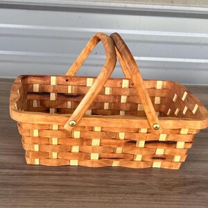 May include: A rectangular woven basket constructed from alternating light and medium brown wooden strips. The basket features two arched wooden handles attached to the sides with small brass-colored metal screws, creating a sturdy open-top container.