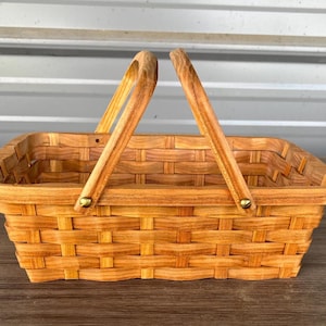May include: A rectangular, woven wooden basket with two arched handles. The basket is made of light brown wood and has a woven pattern. The handles are attached with brass-colored hardware. The basket is empty.