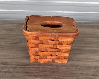 Könnte beinhalten: Ein quadratischer Weidenkorb mit Holzdeckel. Der Korb besteht aus hellbraunem, gewebtem Material und hat eine rechteckige Öffnung in der Mitte des Deckels. Der Korb steht auf einer Holzoberfläche.