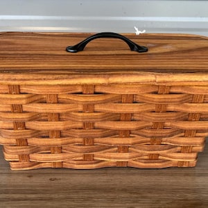 May include: A rectangular woven basket with a light brown wood grain finish and a matching flat wooden lid. The lid features a single curved black metal handle centered on top. The basket has a structured, woven design with horizontal slats.