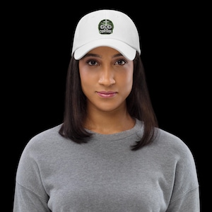 Könnte beinhalten: Eine weiße Baseballkappe mit einer kreisförmigen Grafik auf der Vorderseite. Die Grafik zeigt einen grünen, strukturierten Kranz, der ein kleines Kreuz und den weißen Text "GOD IS IN CONTROL" in der Mitte umschließt.