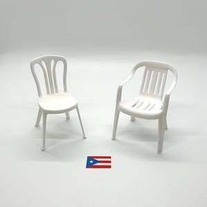 May include: Two miniature white plastic chairs, one armless with a vertical slat back and one with armrests and a slatted back, positioned on a plain surface with a small rectangular sticker featuring the Puerto Rican flag centered between them.