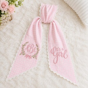 May include: A light pink and white gingham fabric hair ribbon featuring a scalloped white lace edge. The left side displays an embroidered floral wreath surrounding the letters MBA, while the right side features the embroidered text It's a Girl.