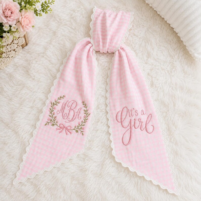May include: A light pink and white gingham fabric hair ribbon featuring a scalloped white lace edge. The left side displays an embroidered floral wreath surrounding the letters MBA, while the right side features the embroidered text It's a Girl.