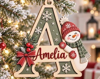May include: A wooden letter A Christmas ornament featuring a green background with white snowflake cutouts, a red bow, green pine needles, red berries, and a snowman wearing a red hat and green scarf with the name Amelia in red script.