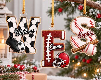 May include: Three personalized acrylic letter ornaments hanging from gold cords. The letter W features a soccer ball pattern with a black cleat, the letter E has a textured football design with a red helmet, and the letter S displays a baseball pattern with a bat and ball.