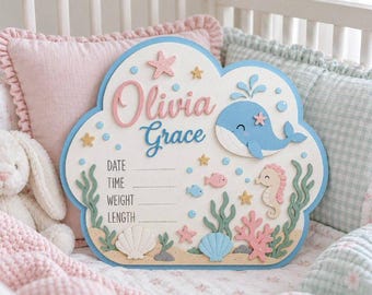 May include: A cloud-shaped wooden birth announcement sign featuring the name Olivia Grace in pink and blue script. The board displays a blue whale, seahorse, seashells, starfish, and seaweed illustrations with blank lines for date, time, weight, and length.
