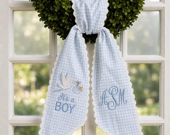 May include: A light blue and white gingham fabric sash with scalloped edges. The left side features an embroidered stork carrying a baby and the text It's a BOY. The right side features an embroidered monogram with the letters ASM in blue thread.