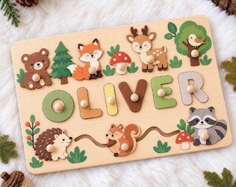 May include: A rectangular wooden name puzzle featuring the name OLIVER in colorful block letters with small knobs. The board is decorated with illustrations of a bear, fox, deer, bird, hedgehog, squirrel, raccoon, trees, and mushrooms.