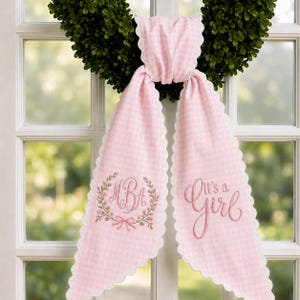 May include: A pink and white gingham fabric sash with scalloped edges, draped over a green wreath. The left side features an embroidered floral wreath surrounding the monogram MBA, while the right side displays the embroidered text It's a Girl.