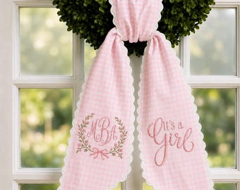 May include: A pink and white gingham fabric sash with scalloped edges, draped over a green wreath. The left side features an embroidered floral wreath surrounding the monogram MBA, while the right side displays the embroidered text It's a Girl.