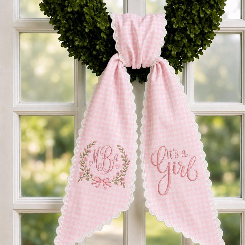 May include: A pink and white gingham fabric sash with scalloped edges, draped over a green wreath. The left side features an embroidered floral wreath surrounding the monogram MBA, while the right side displays the embroidered text It's a Girl.