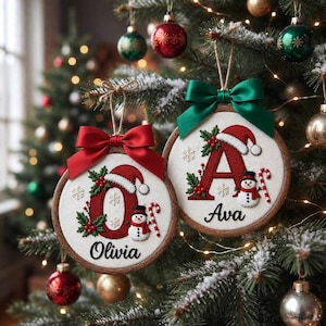 Könnte beinhalten: Zwei runde Holzstickrahmen-Ornamente an einem Weihnachtsbaum. Eines zeigt ein rotes O mit Weihnachtsmütze, Stechpalme und Schneemann, beschriftet mit Olivia. Das andere hat ein rotes A mit Weihnachtsmütze und Schneemann, beschriftet mit Ava.
