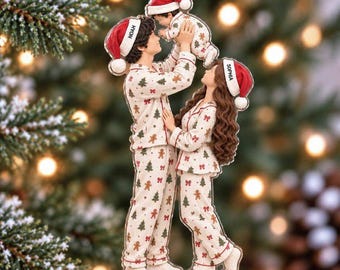 May include: A flat acrylic Christmas ornament featuring a family of three wearing matching cream-colored pajamas with gingerbread men and tree patterns. The figures wear red Santa hats labeled Noah, Sophia, and a baby, with the parent lifting the baby.