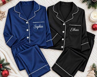May include: Two sets of long-sleeved button-down pajamas, one in navy blue and one in black, both featuring white piping trim. The navy set is embroidered with the name Sophia, and the black set is embroidered with the name Ethan.