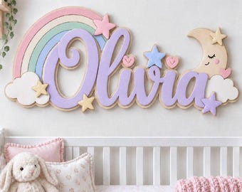 May include: A wall-mounted wooden name sign featuring the name Olivia in large purple script letters. The design includes a pastel rainbow, white clouds, a smiling crescent moon, and several small stars and hearts in pink, blue, and yellow.