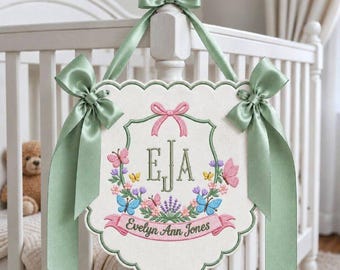 May include: A scalloped, off-white fabric wall hanging featuring embroidered floral motifs, colorful butterflies, and the monogram EJA. A pink banner displays the name Evelyn Ann Jones. The piece is accented with sage green satin ribbons.