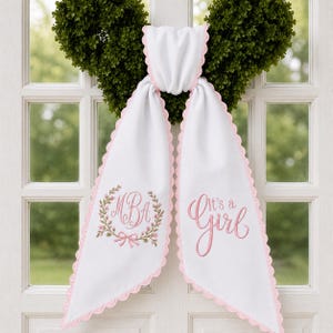 May include: A white fabric door hanger with scalloped pink edges. The left side features an embroidered floral wreath surrounding the monogram MBA in pink. The right side features the embroidered pink text It's a Girl in a cursive script font.