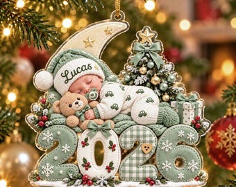 May include: A Christmas ornament featuring a sleeping baby wearing a green hat with the name Lucas, holding a teddy bear. The design includes a crescent moon, a decorated Christmas tree, a gift box, and the year 2026 in large green numbers.