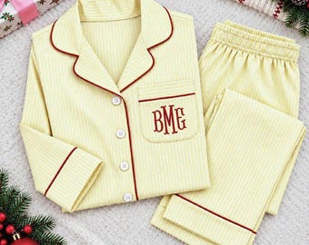 May include: A light yellow pajama set with thin white vertical stripes and red piping trim. The long-sleeved button-down shirt features a chest pocket embroidered with the red monogram letters BMG. The matching pants have an elastic waistband.
