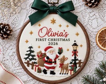 May include: A round wooden embroidery hoop featuring white fabric with the embroidered text Olivia's FIRST CHRISTMAS 2026. The design includes a Santa Claus figure, a reindeer, a Christmas tree, a lamp post, and gifts, topped with a dark green bow.