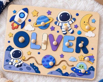May include: A wooden peg puzzle featuring the name OLIVER in large, colorful letters. The board is decorated with space-themed illustrations including astronauts, a rocket ship, planets, a crescent moon, and stars in shades of blue, yellow, and purple.