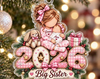 May include: A clear acrylic Christmas ornament featuring a girl holding a baby, both wearing pink gingham pajamas. The design includes a stuffed bunny, a gift box, the year 2026, and the text 'Emma', 'Ava', and 'Big Sister'.