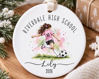 May include: A round white ceramic ornament featuring a watercolor illustration of a soccer player in a pink jersey with the number 10, kicking a ball. The text reads Riverdale High School, the name Lily, and the year 2026 in black script.