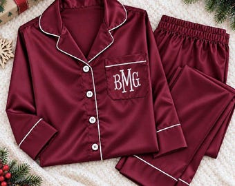 May include: A burgundy satin pajama set featuring a long-sleeved button-down shirt with white piping and a chest pocket embroidered with the white monogram letters BMG, paired with matching long pants with an elasticized waistband.