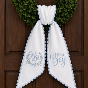 May include: A white fabric door hanger with light blue scalloped edges. The left side features an embroidered wreath with the monogram MBA and a small bow. The right side displays the embroidered blue text It's a Boy in a script font.