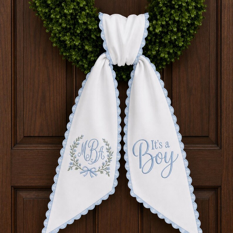 May include: A white fabric door hanger with light blue scalloped edges. The left side features an embroidered wreath with the monogram MBA and a small bow. The right side displays the embroidered blue text It's a Boy in a script font.