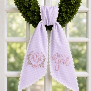 May include: A lavender and white gingham fabric sash with scalloped white edges. The left side features an embroidered floral wreath surrounding the letters MBA, while the right side displays the embroidered pink text It's a Girl.