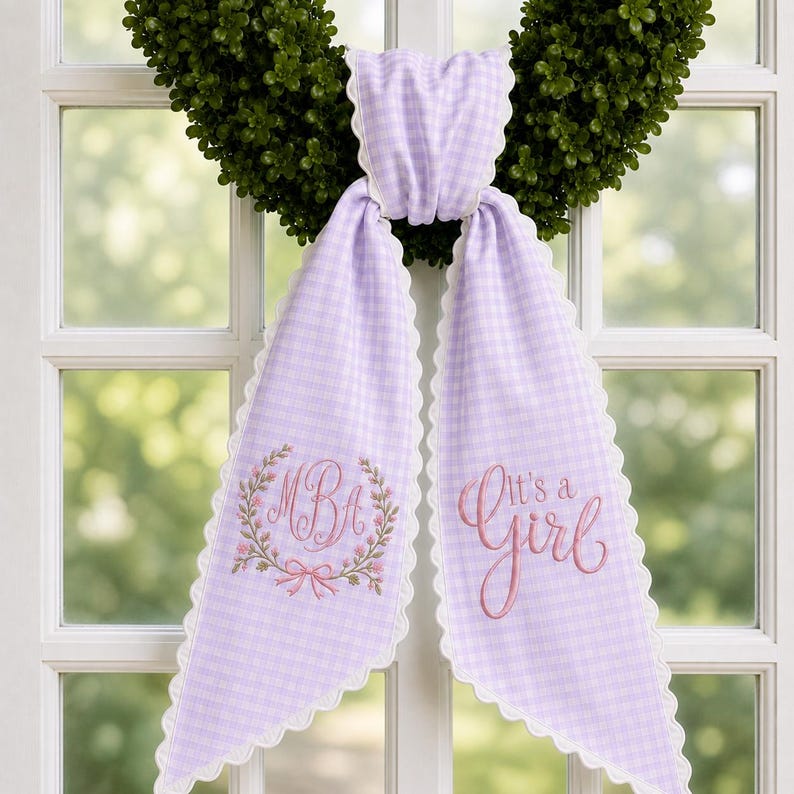 May include: A lavender and white gingham fabric sash with scalloped white edges. The left side features an embroidered floral wreath surrounding the letters MBA, while the right side displays the embroidered pink text It's a Girl.