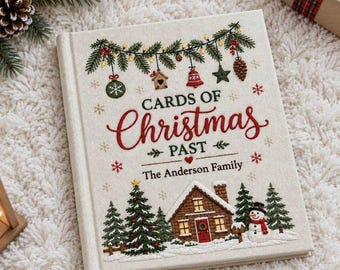May include: A cream-colored hardcover book titled Cards of Christmas Past The Anderson Family, featuring embroidered holiday motifs including pine branches, ornaments, a house, a snowman, and snowflakes in red, green, and brown.