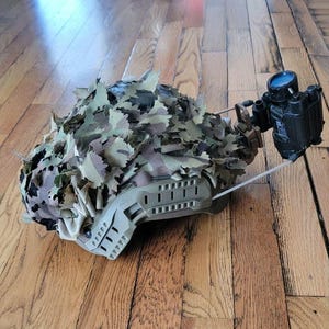 May include: A tactical helmet covered in a camouflage leaf-patterned fabric cover, featuring a tan side rail system and a black night vision monocular device mounted to the front with a thin wire cable extending from the unit.