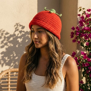 May include: A bright red crocheted beanie hat featuring a rolled brim and a small, light green crocheted stem and leaf detail attached to the top centre. The hat is constructed with a dense, textured yarn stitch pattern throughout the entire piece.
