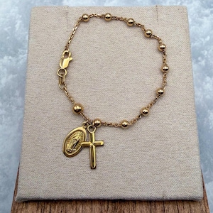 Op de afbeelding: Een goudkleurige kettingarmband met kleine, gelijkmatig verdeelde bolvormige kralen. De armband is voorzien van een karabijnsluiting en twee bungelende bedels: een kleine ovale hanger met een figuur en een eenvoudig, glad kruisje.