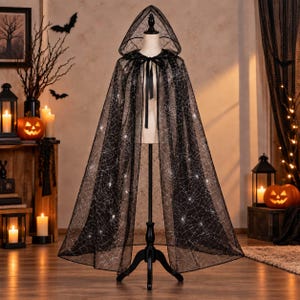 May include: A floor-length black hooded cape made of sheer mesh fabric featuring a white spiderweb pattern throughout. The garment includes a black ribbon tie closure at the neck and is displayed on a mannequin stand, showcasing a wide silhouette.